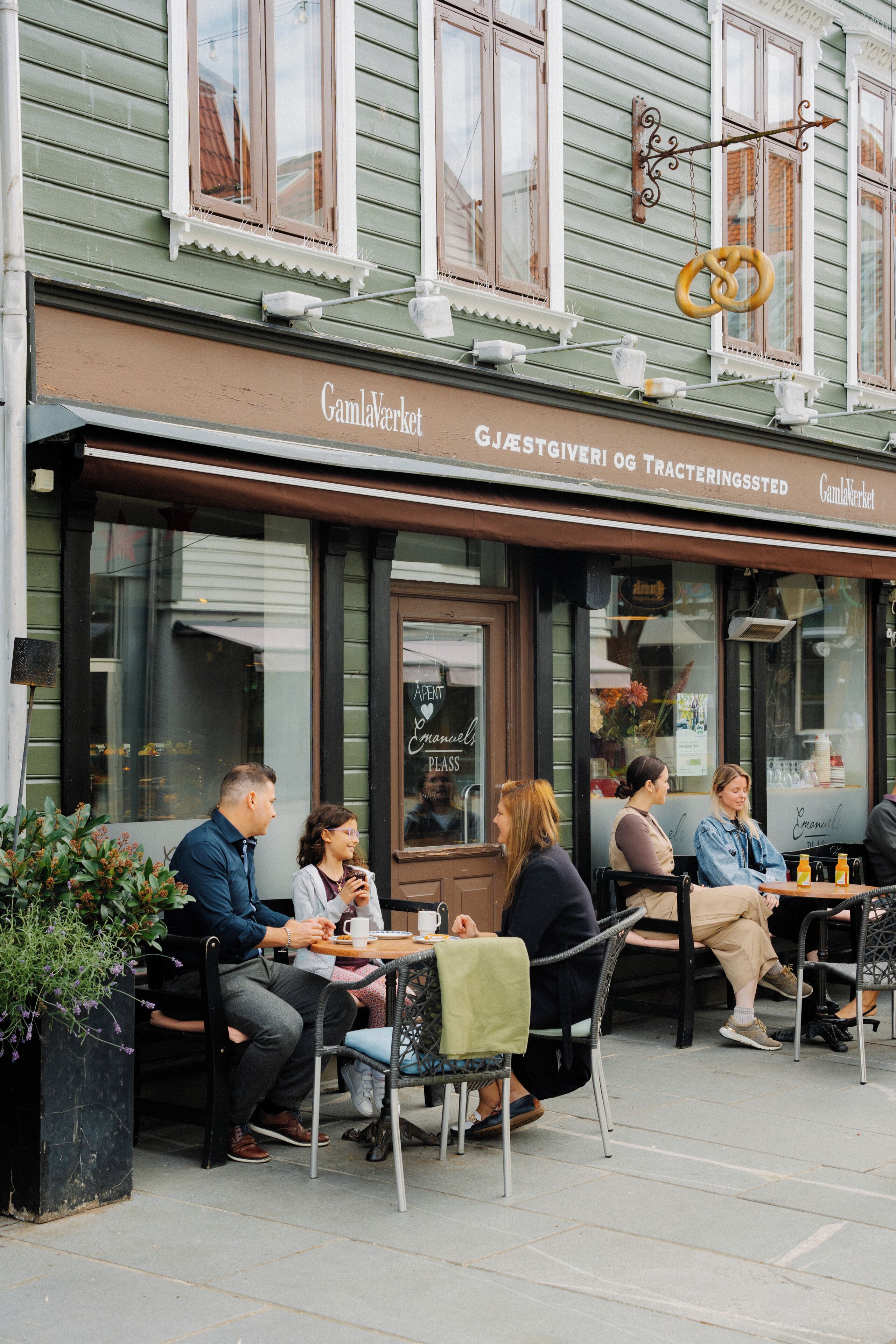Un café en plein air avec des tables et des chaises, une enseigne indiquant « Gjestegiver og Trakteringssted » et un bretzel suspendu au-dessus.