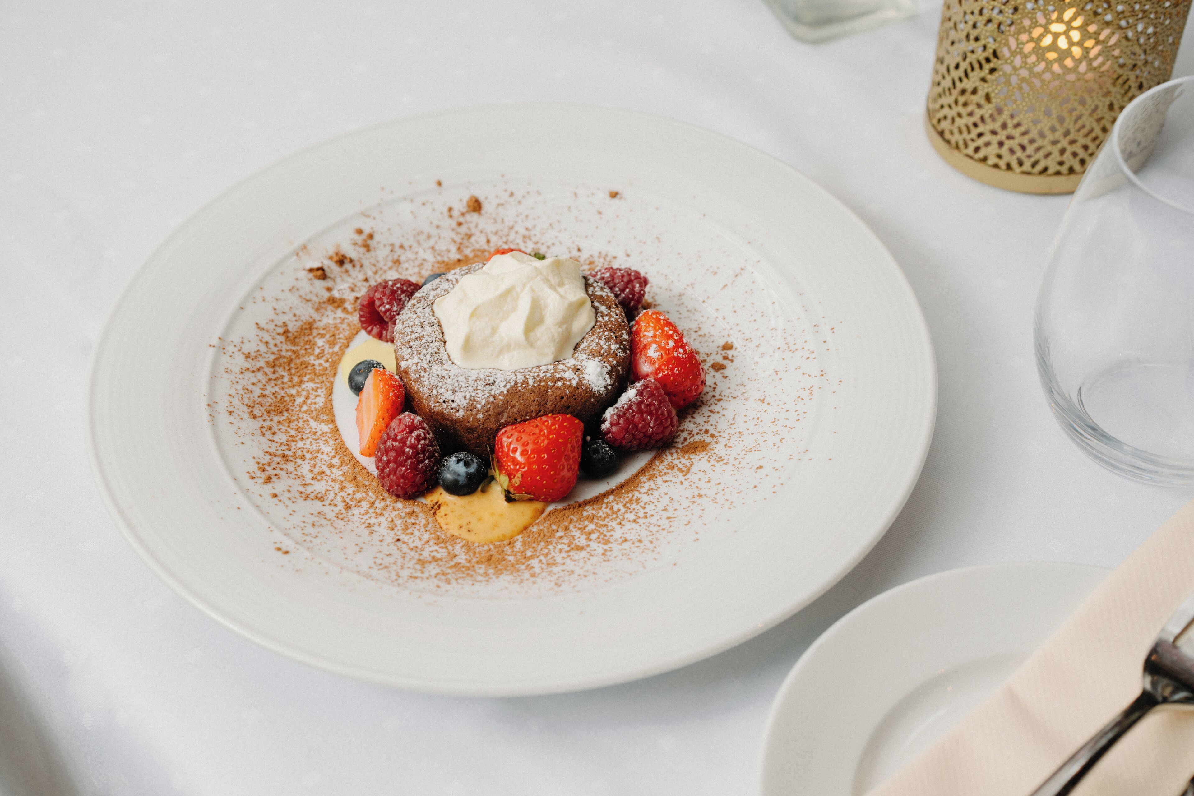 Un postre en un plato blanco con fresas, frambuesas y una cucharada de crema, espolvoreado con azúcar glas.
