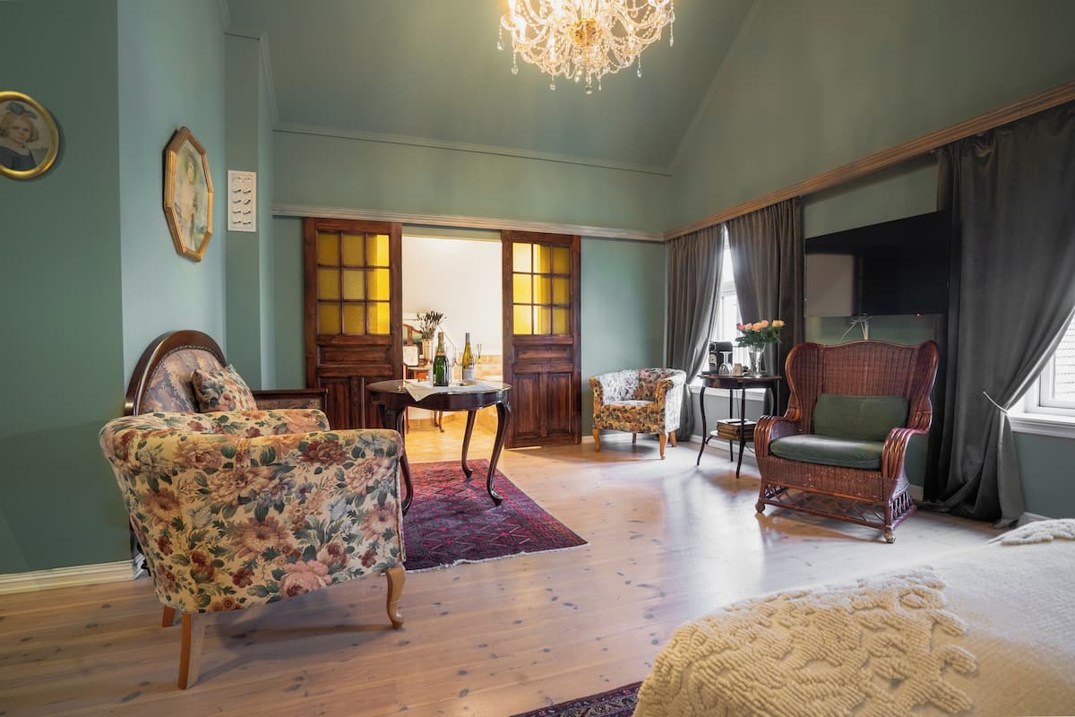 Um quarto espaçoso com teto alto, um candelabro, poltronas com padrão floral, uma cadeira de vime e piso de madeira.