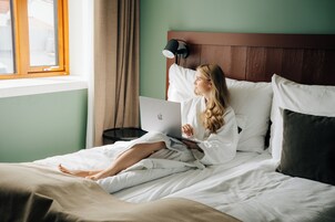 Una persona acostada en una cama con un ordenador portátil, una lámpara de noche y una ventana con cortinas.