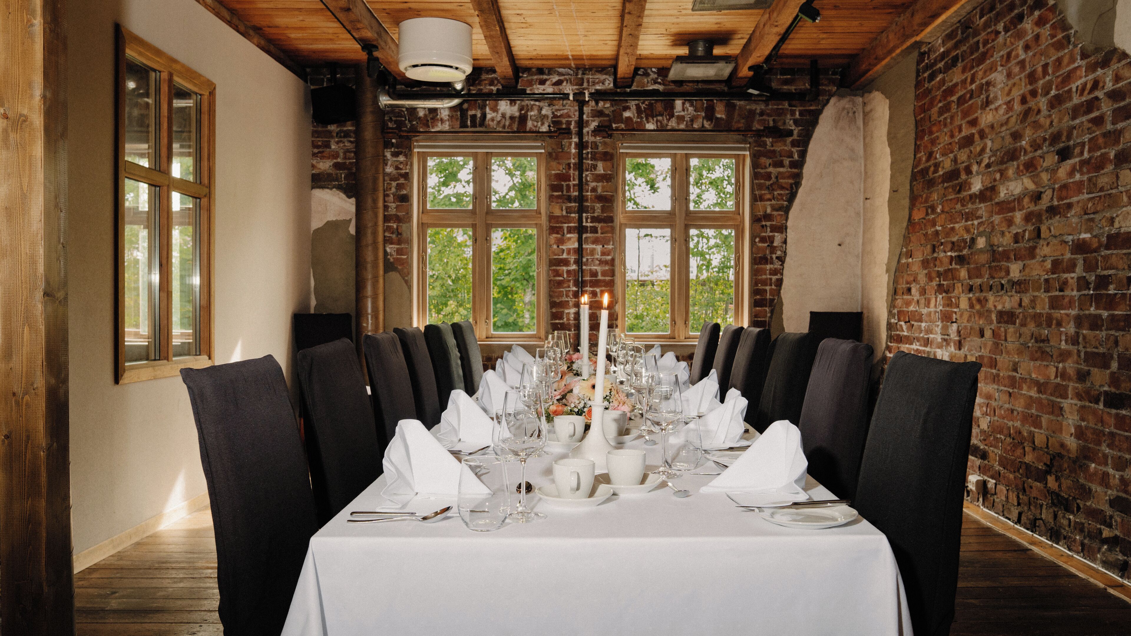 A dining room with a long table set for a meal, featuring a brick wall and large windows.