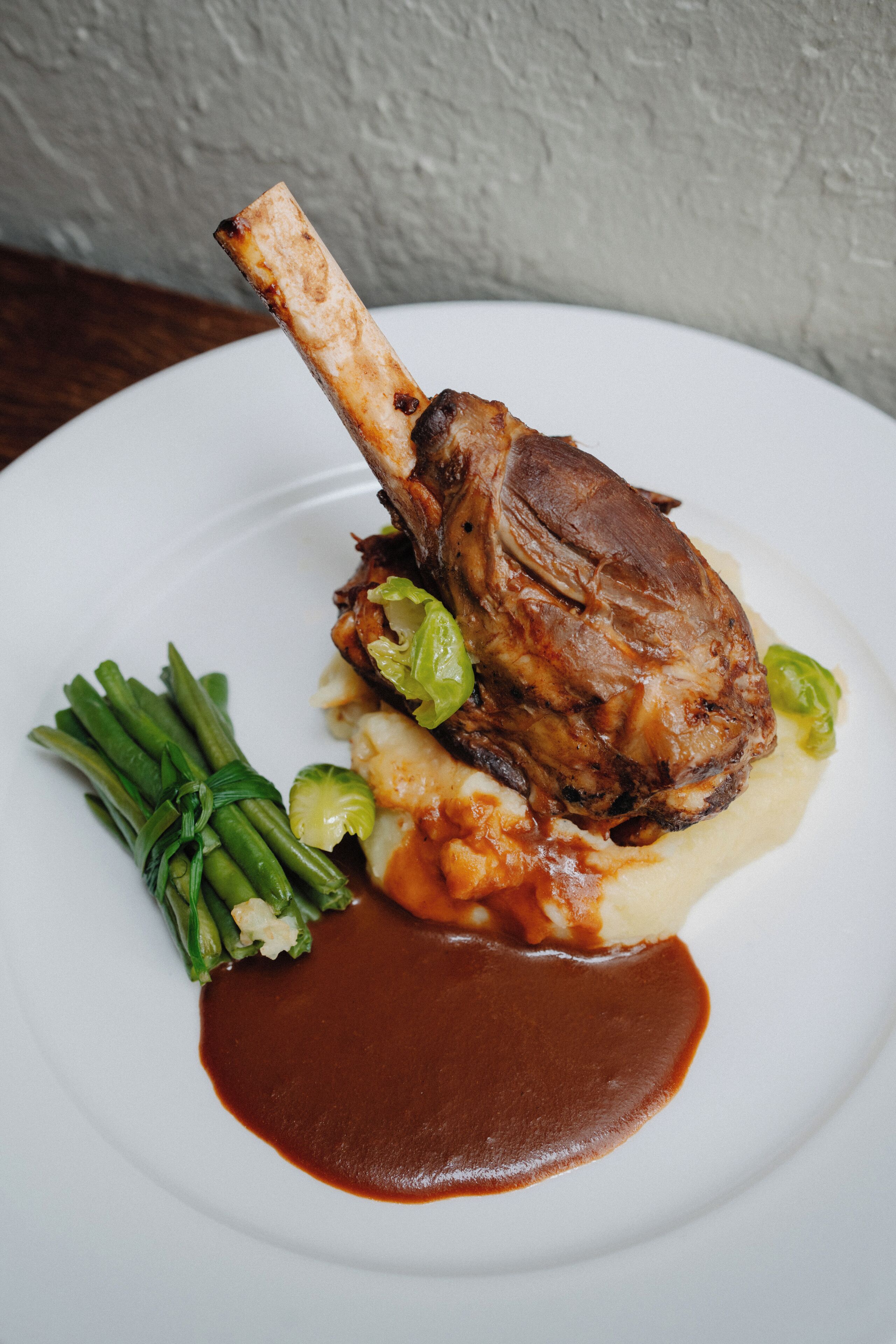 A plate with a lamb shank, green beans, and mashed potatoes.