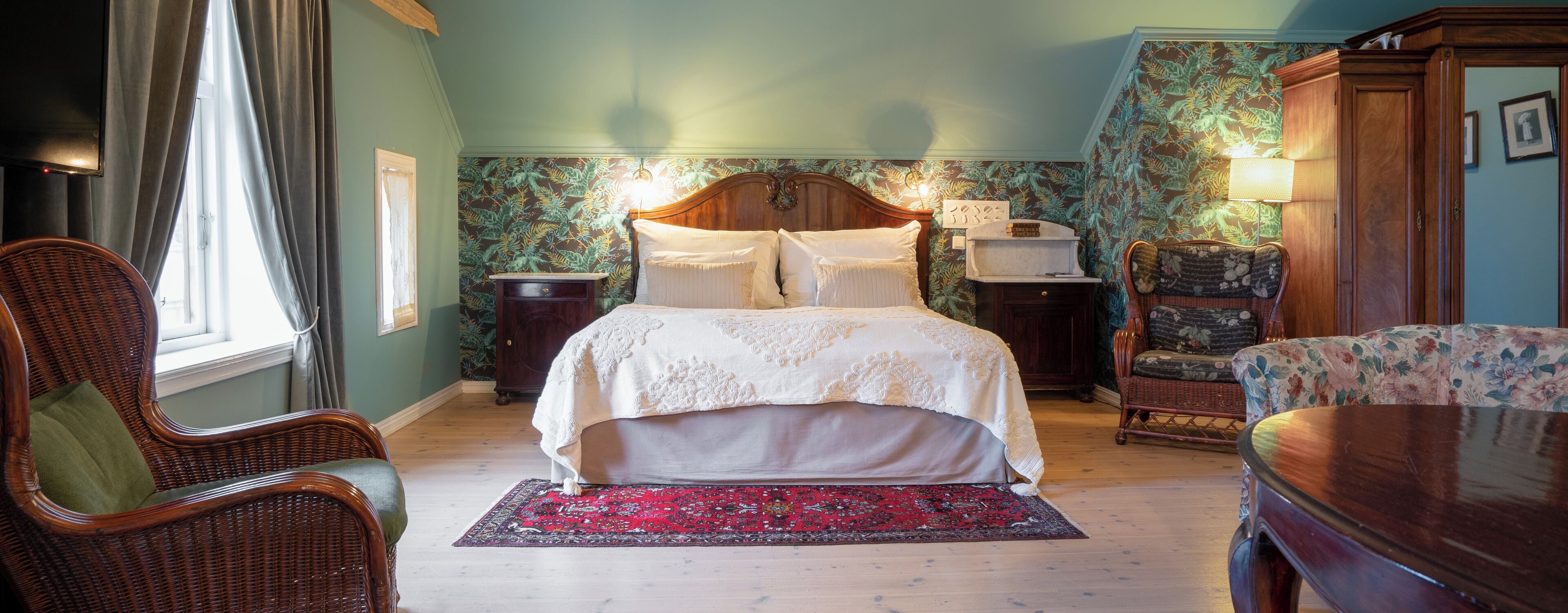 Un dormitorio con una cama grande, una mesita de noche de madera, una silla de mimbre, una mesa de madera y un tapete estampado.