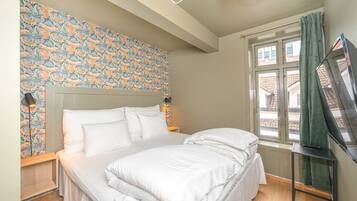 Un dormitorio con una cama grande, papel tapiz estampado, una ventana con cortinas y un televisor sobre un soporte.