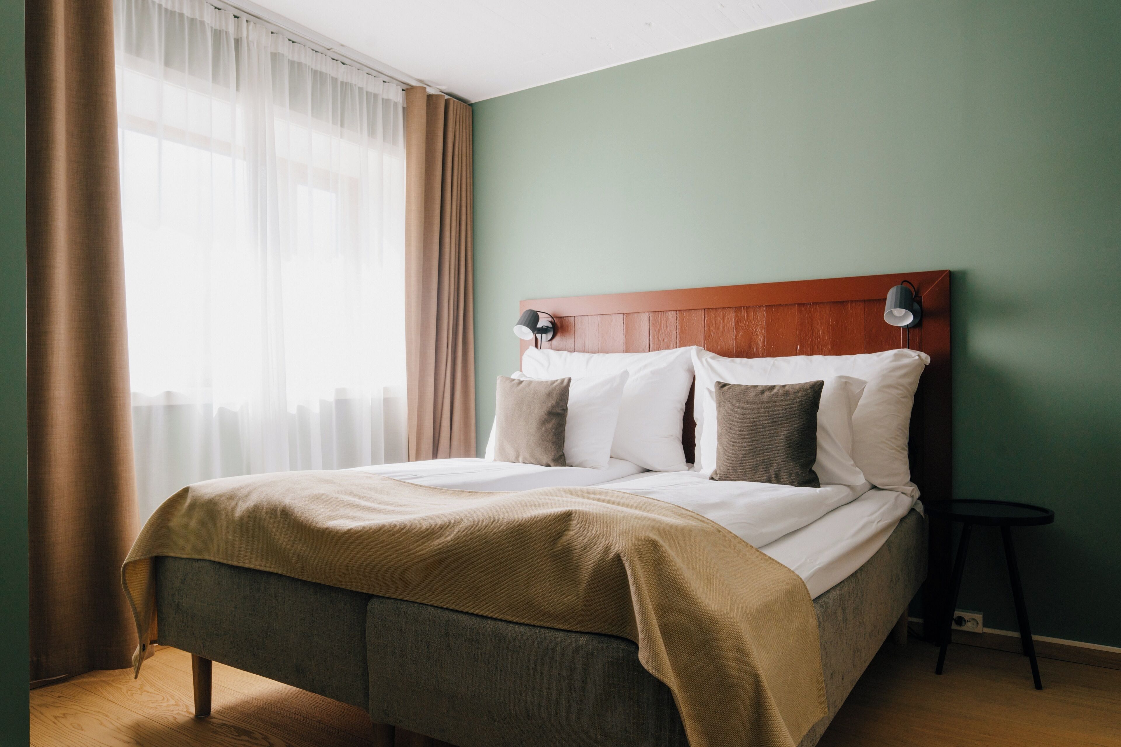 A neatly made bed with a wooden headboard, a bedside table with a lamp, and a window with sheer curtains.