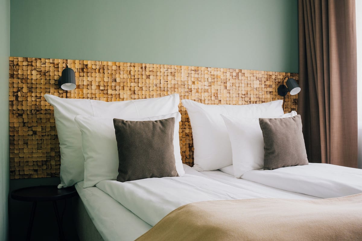 Cama bem arrumada com almofadas brancas e castanhas, cabeceira de tecido e duas luminárias fixadas na parede.