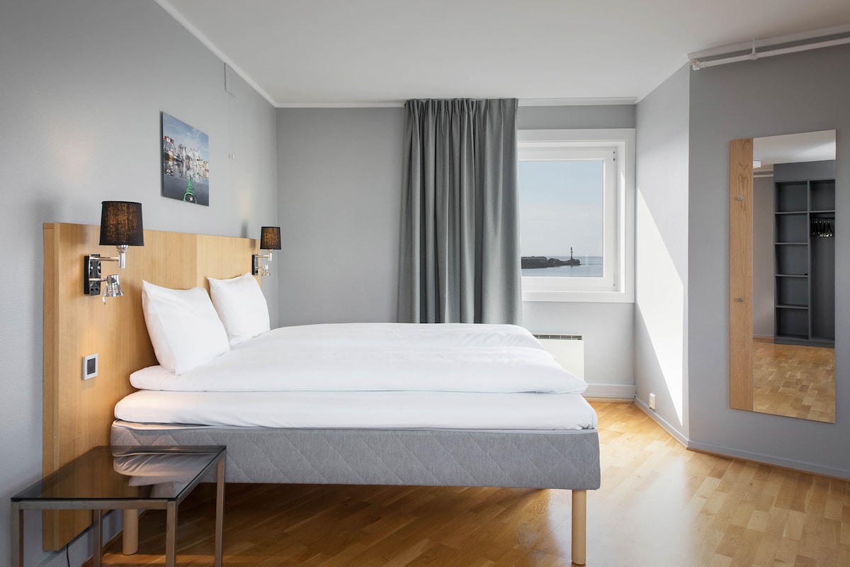 A modern hotel room with a large bed, a bedside table, a mirror, and a window with a view.