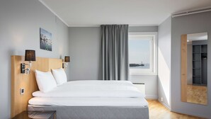 A modern hotel room with a large bed, a bedside table, a mirror, and a view of the sea through the window.