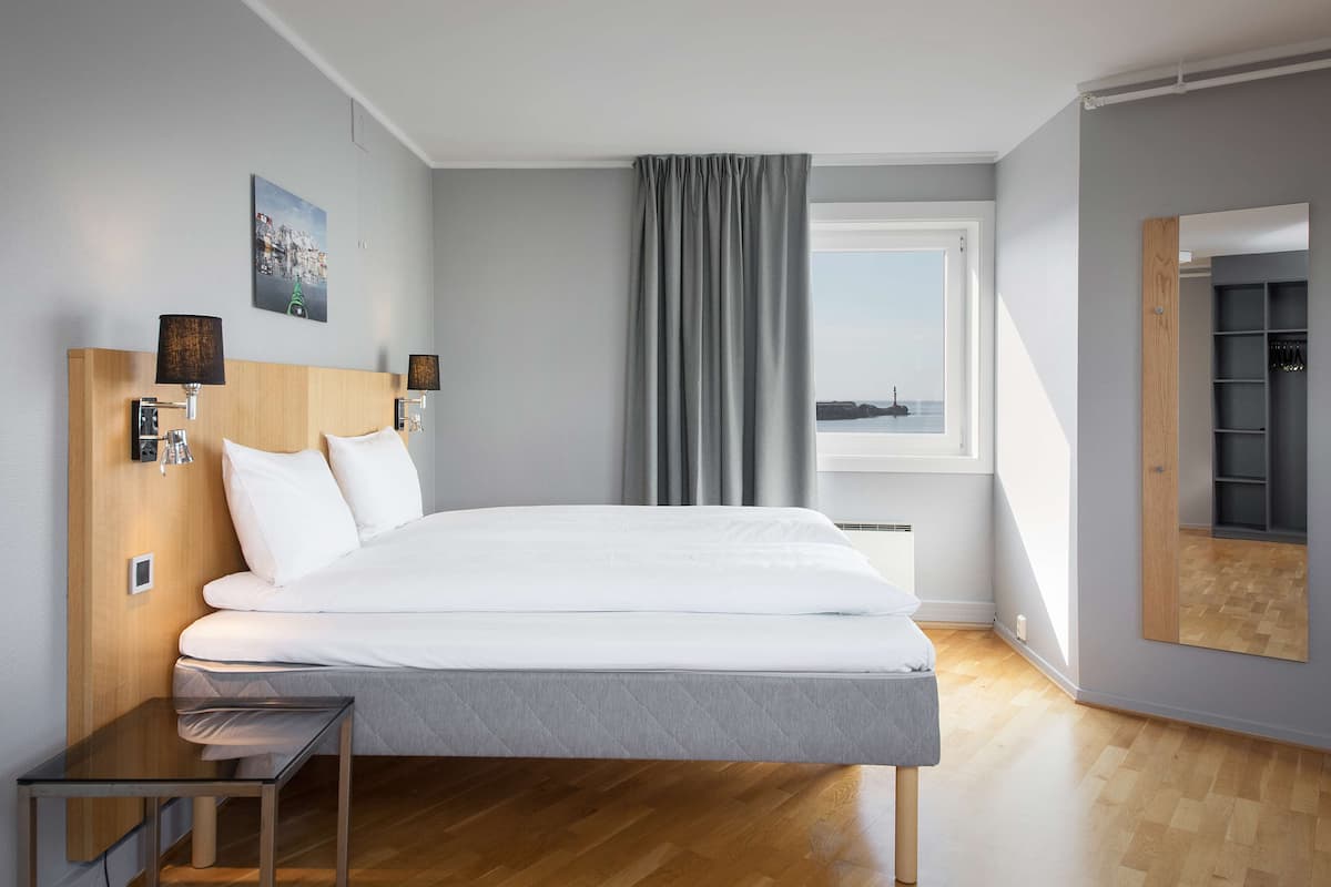 Une chambre d’hôtel moderne avec un grand lit, une table de chevet, un miroir et une vue sur la mer par la fenêtre.
