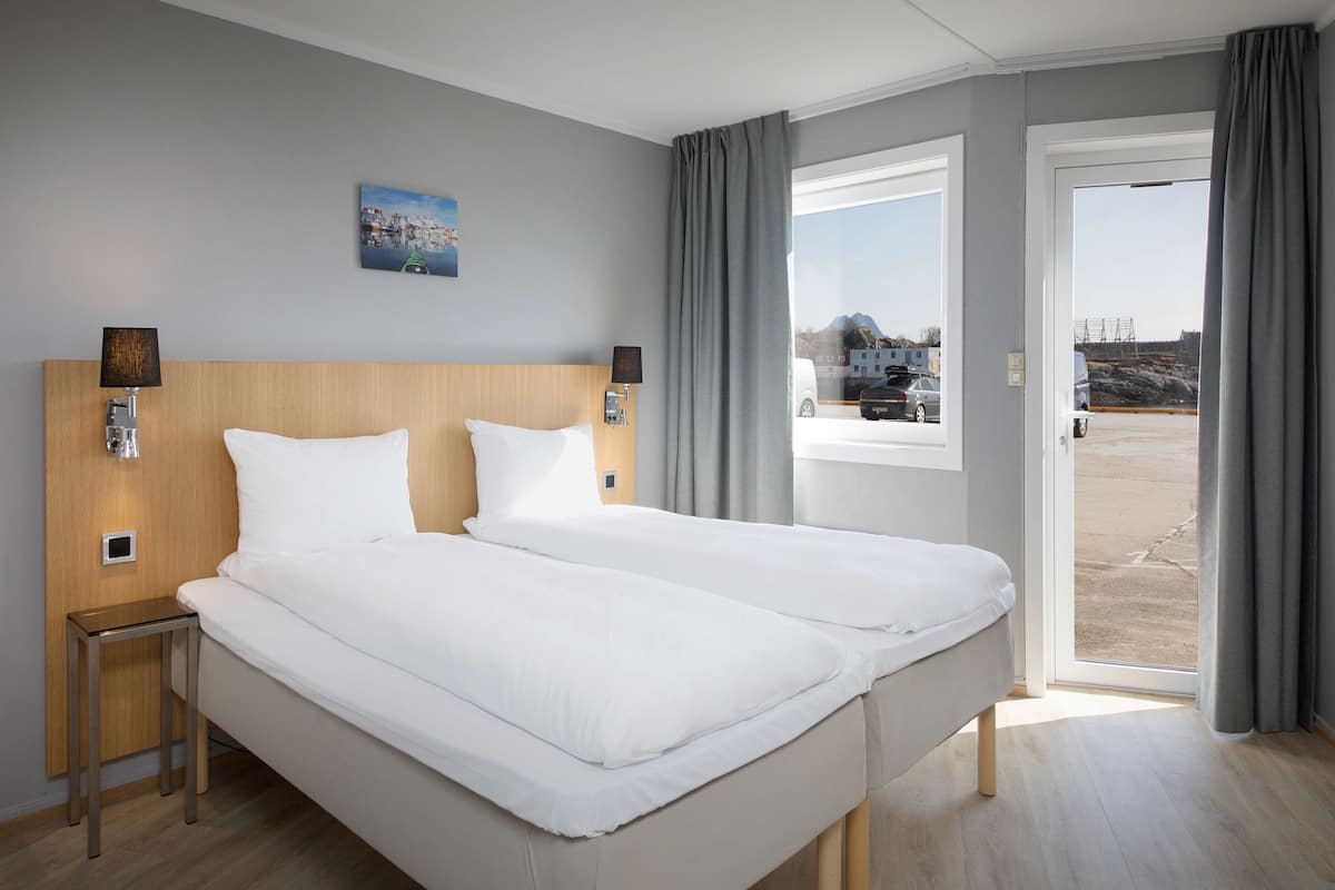 Un lit double avec du linge de lit blanc, une tête de lit en bois, deux tables de chevet avec des lampes, un tableau au mur et une vue à travers une porte-fenêtre coulissante.