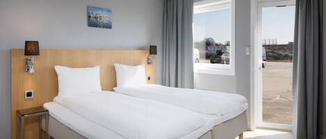 Un lit double avec du linge de lit blanc, une tête de lit en bois, deux tables de chevet avec des lampes, un tableau au mur et une vue à travers une porte-fenêtre coulissante.