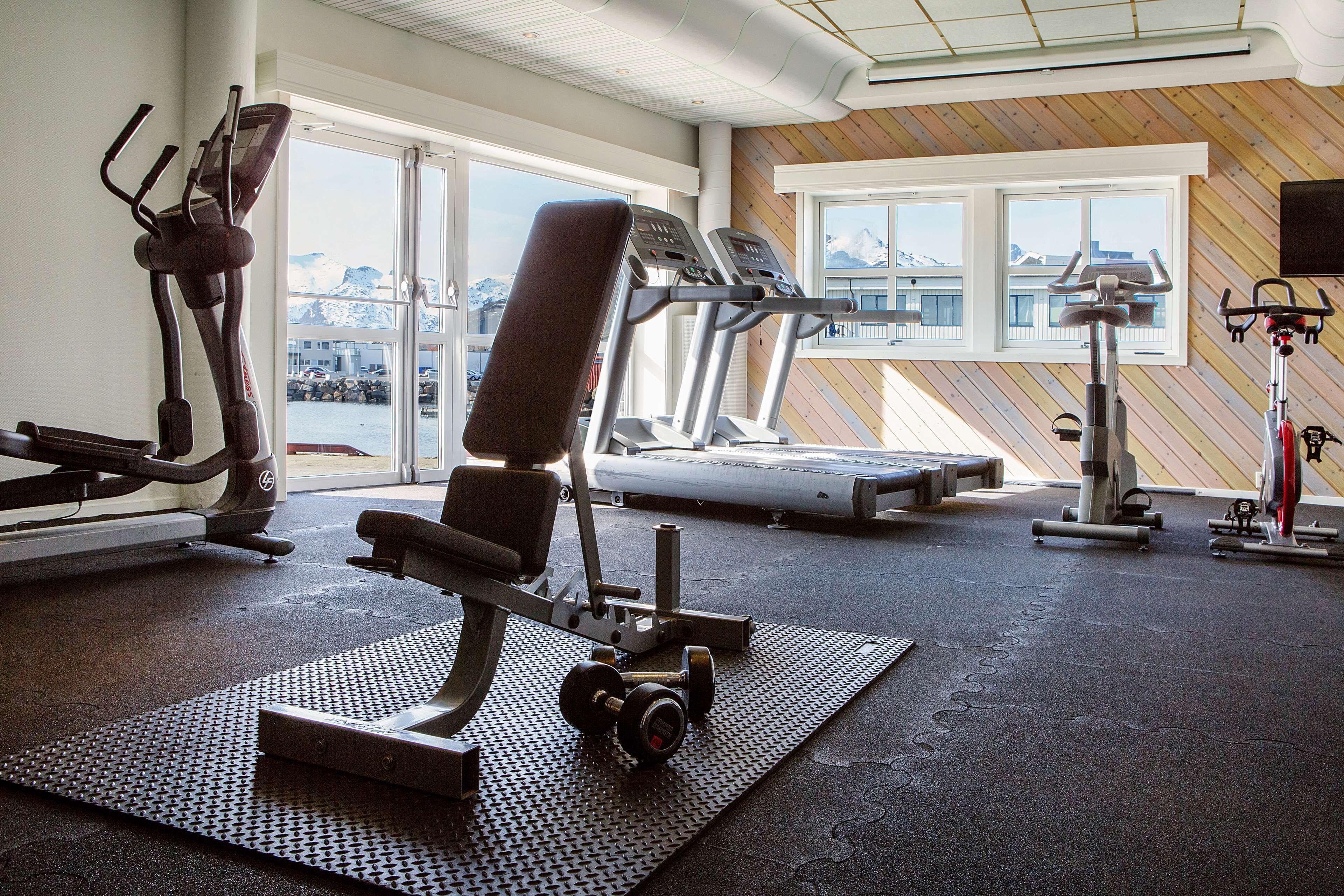 Un gimnasio con equipo de ejercicio, ventanas amplias y vistas a montañas.