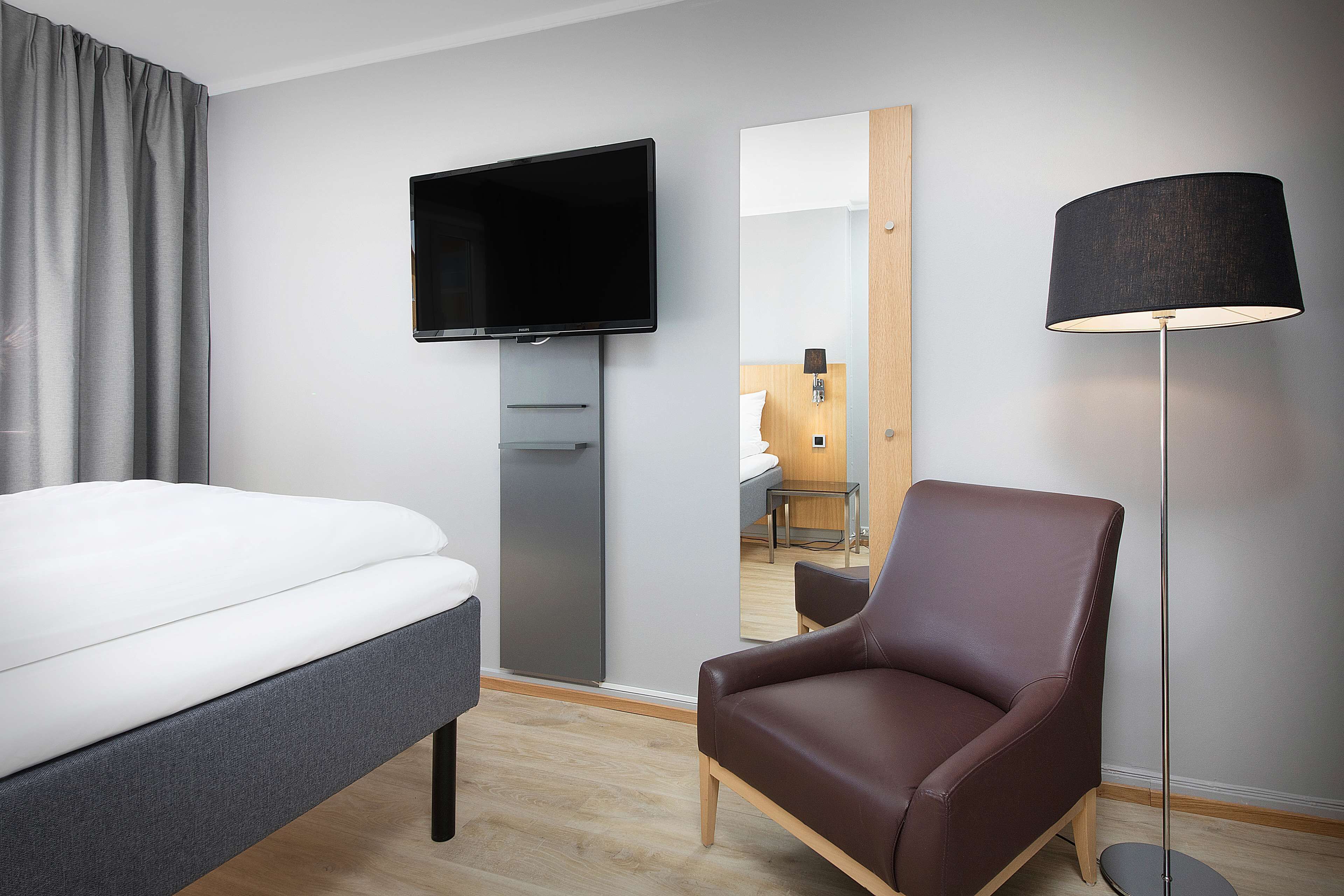 A modern hotel room with a bed, a flat-screen TV, a mirror, a brown leather chair, and a floor lamp.