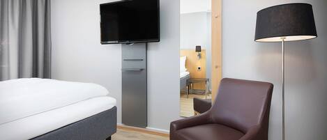 A modern hotel room with a bed, a flat-screen TV, a mirror, a brown leather chair, and a floor lamp.