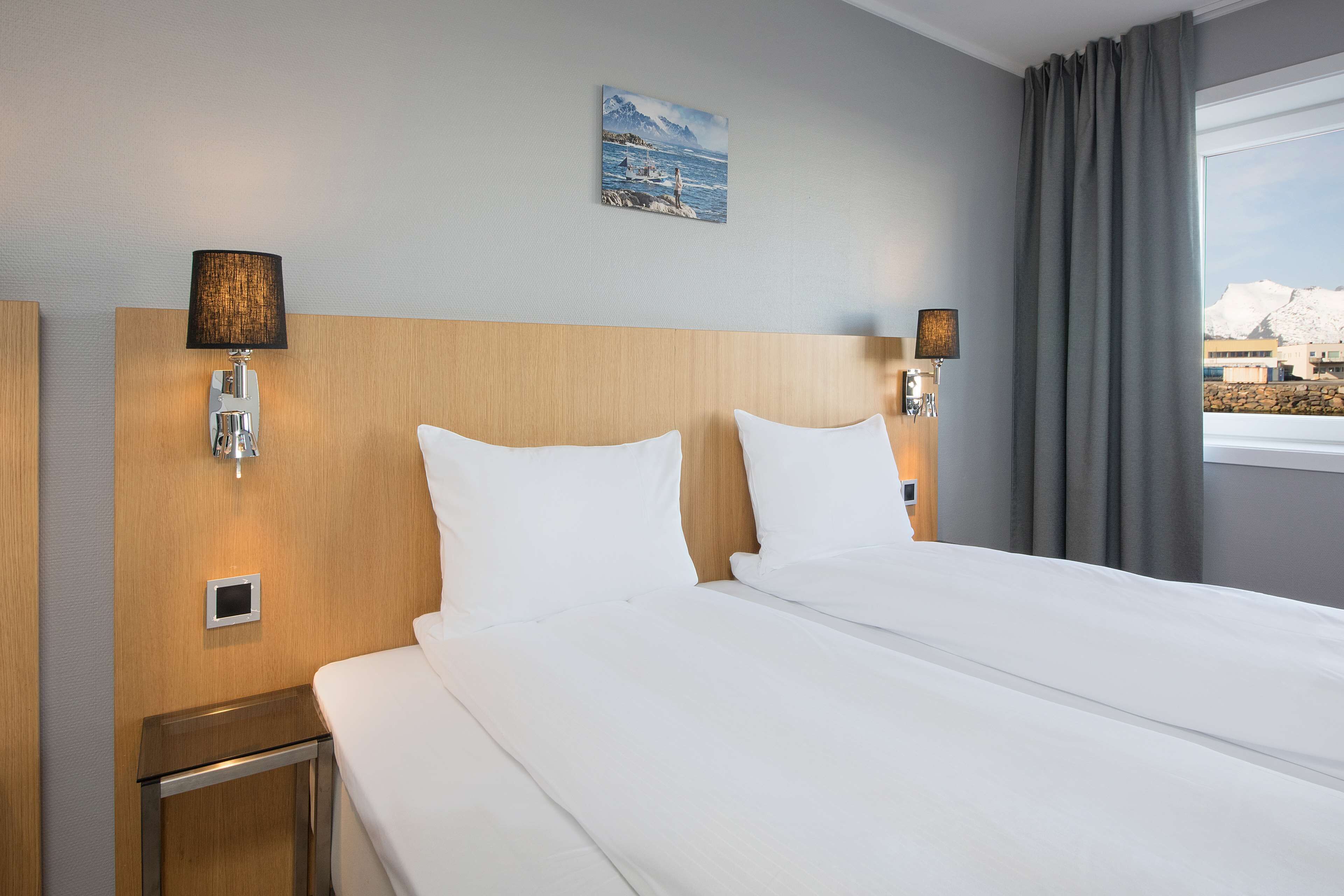 Hotellihuone, jossa on suuri sänky, kaksi yöpöytävalaisinta, pieni pöytä ja ikkuna, jossa on verhot.
