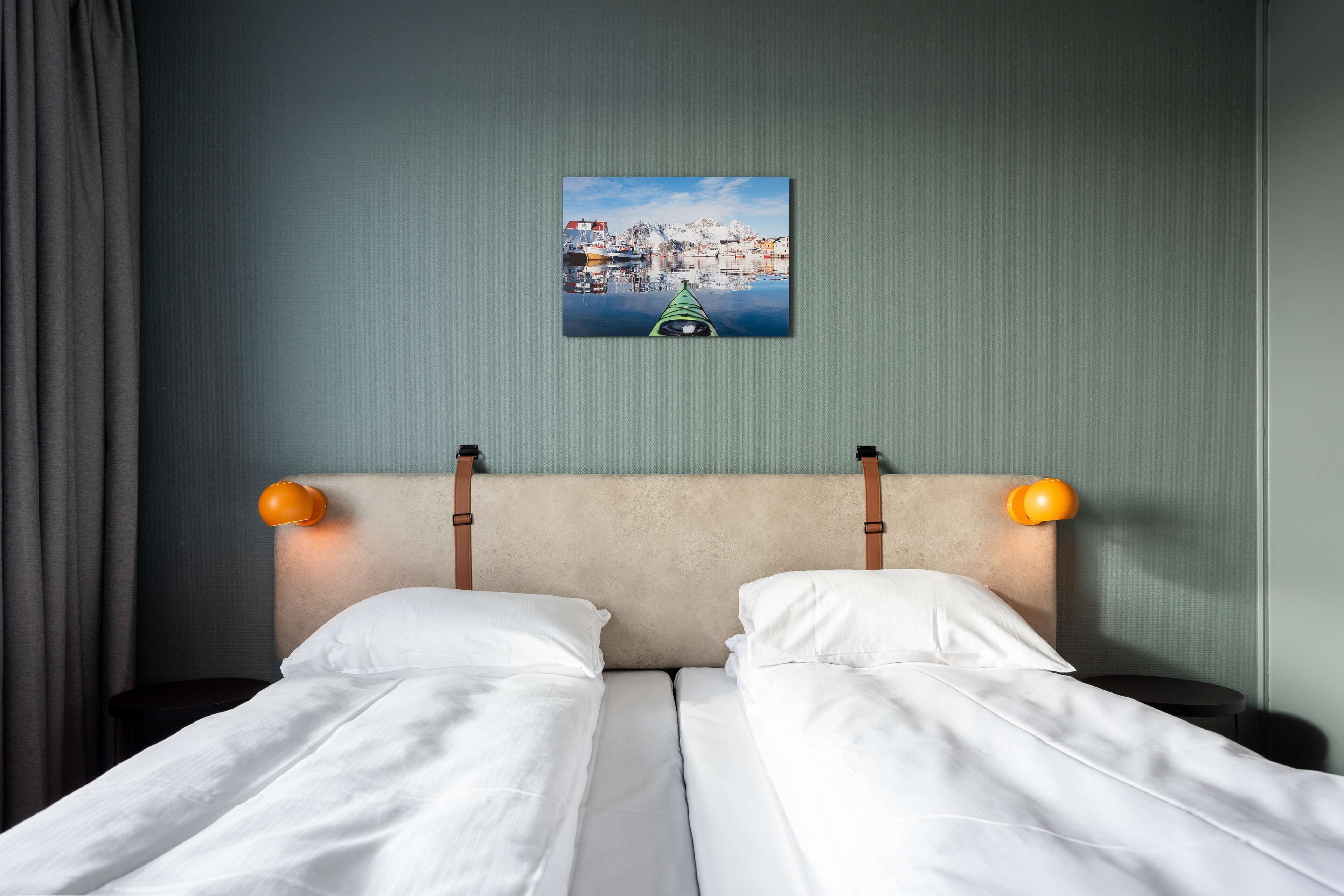 A double bed with white linens, a headboard with orange wall sconces, and a framed picture of a coastal town on the wall.