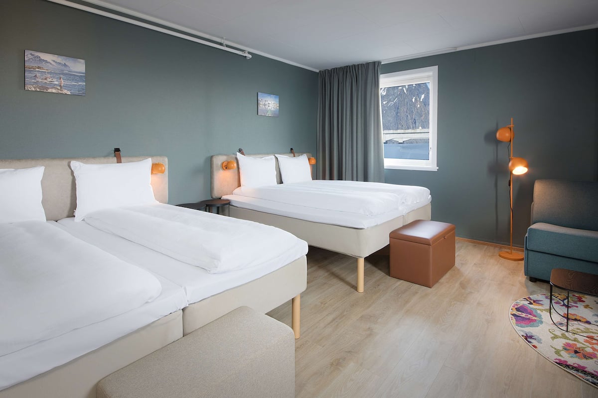 Une chambre d’hôtel avec deux lits, une petite table, une chaise, une lampe et une fenêtre donnant sur les montagnes.