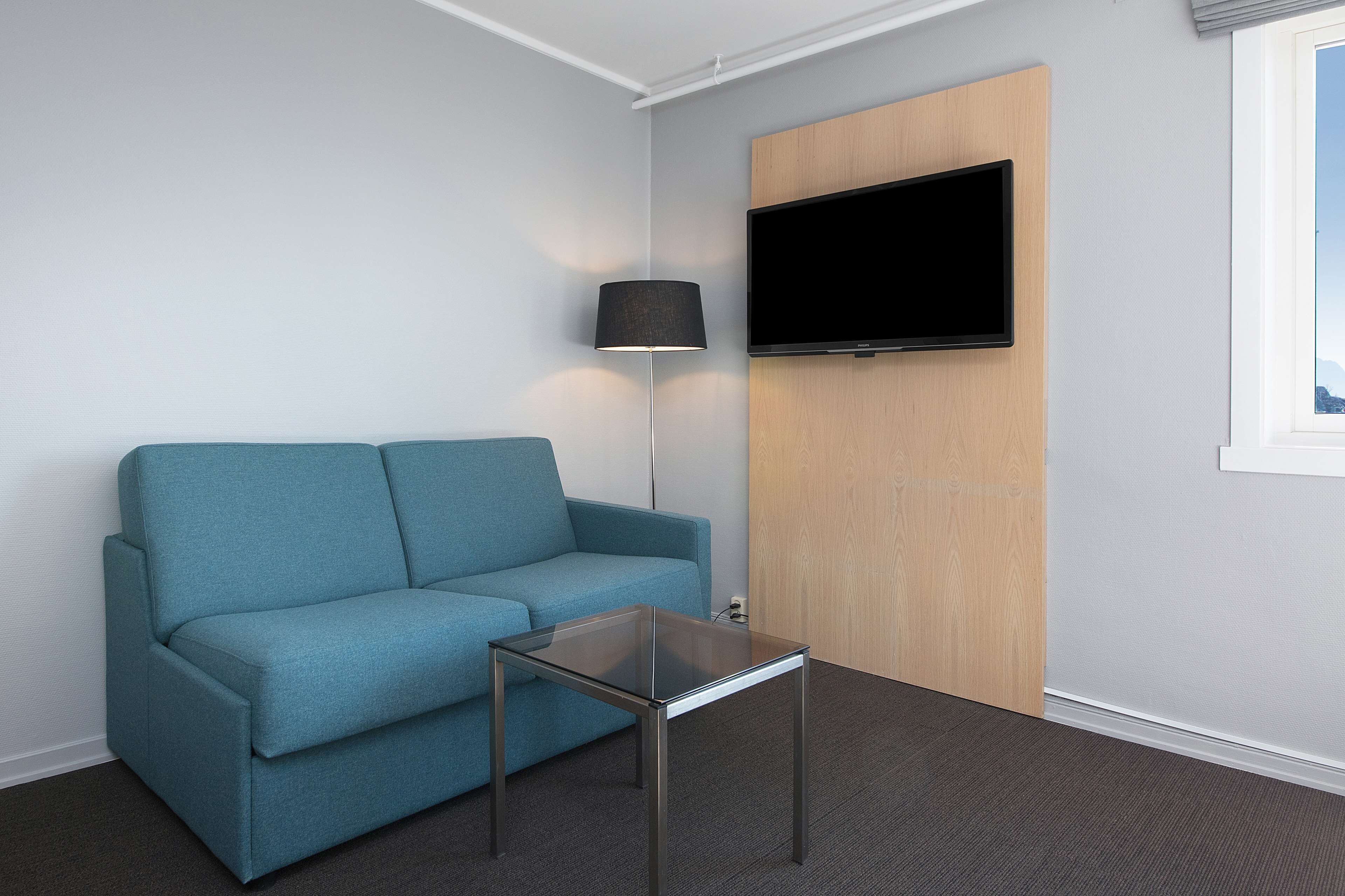 A modern room with a teal sofa, a glass coffee table, a floor lamp, a mounted TV, and a wooden door.