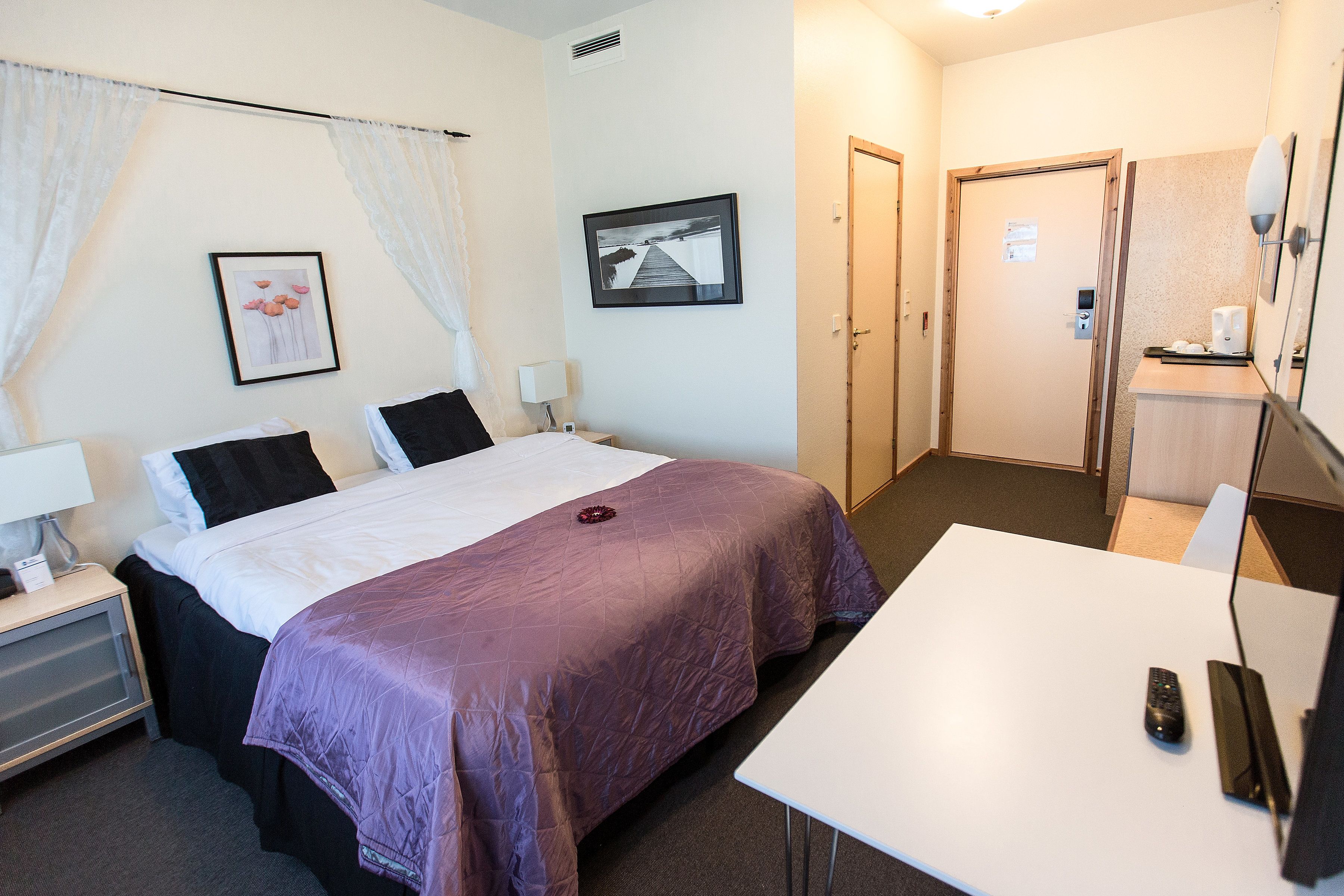 Une chambre d’hôtel avec un lit, des tables de chevet, un bureau et une chaise. Il y a une porte qui donne sur une autre pièce et une fenêtre avec des rideaux.