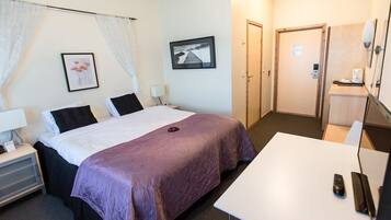 Une chambre d’hôtel avec un lit, des tables de chevet, un bureau et une chaise. Il y a une porte qui donne sur une autre pièce et une fenêtre avec des rideaux.