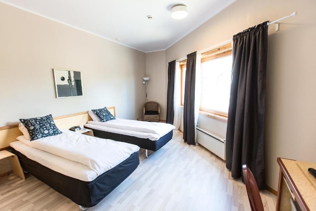 A hotel room with two beds, a bedside table, a lamp, a chair, and a window with curtains.
