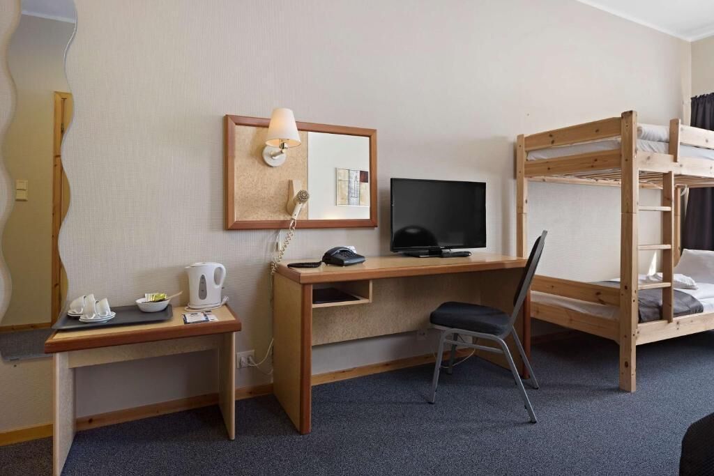 Habitación de hotel con escritorio de madera, una silla, literas, un televisor y una mesita con hervidor y tazas.