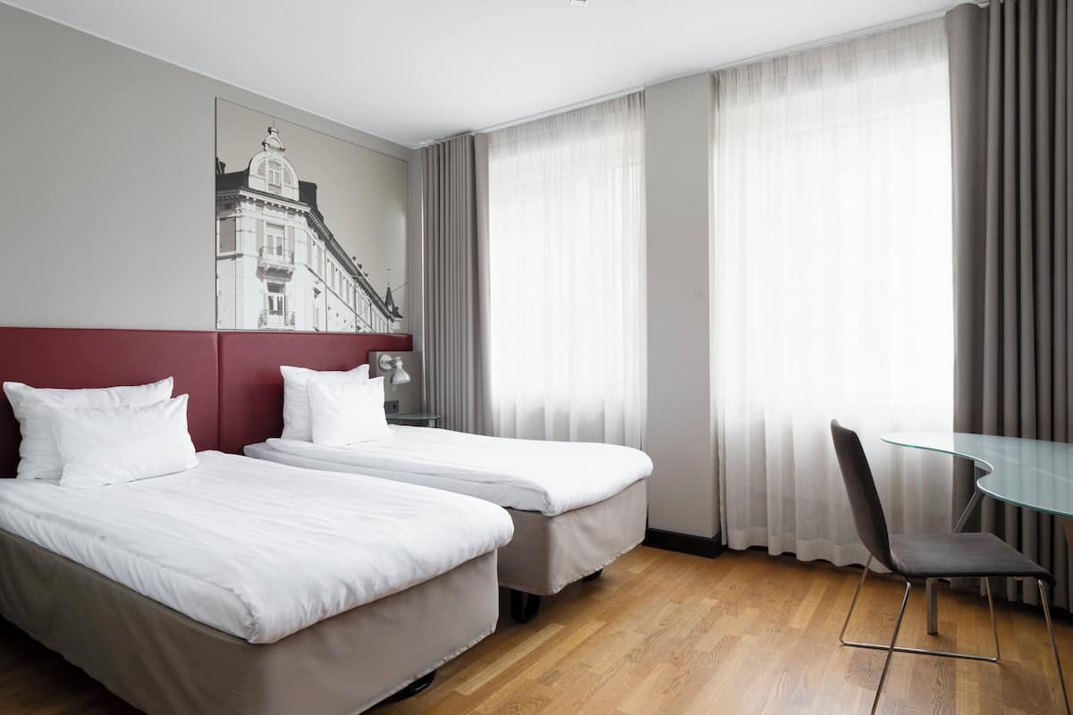 A hotel room with two beds, a glass table, a chair, and a large window with curtains.