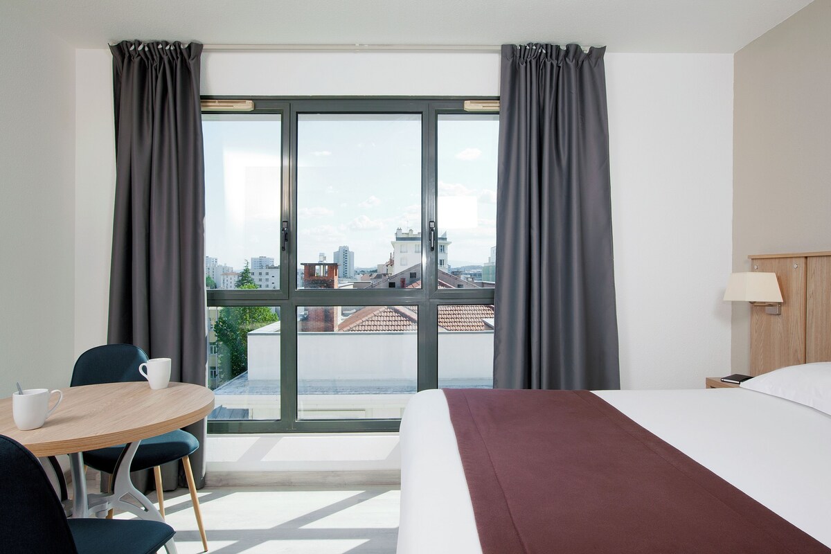 Habitación de hotel con cama, una mesa con silla, ventana con cortinas y vista a edificios.