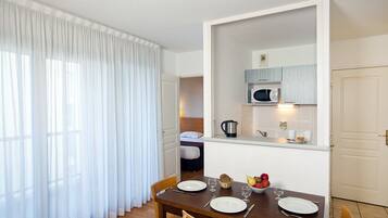 A dining area with a wooden table set for two, a kitchen with a microwave, and a bed in the background.