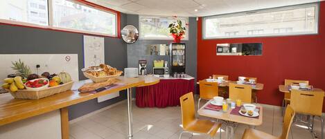 A breakfast area with a long counter, wicker baskets of fruit, and a red tablecloth. There are wooden chairs and tables, and a mirror on the wall.