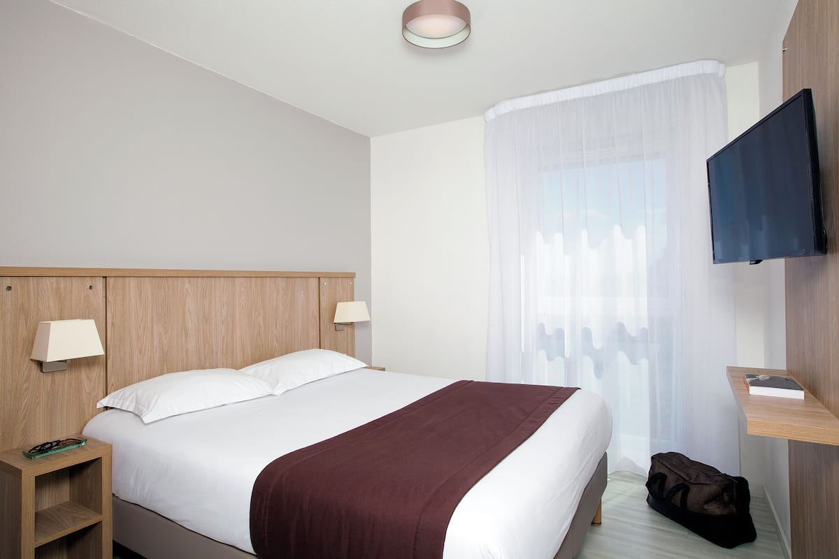 A hotel room with a large bed, a bedside table with a lamp, a TV mounted on the wall, and a window with sheer curtains.