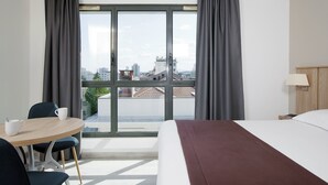 Habitación de hotel con un ventanal grande, una cama, una mesa con sillas y una lámpara.