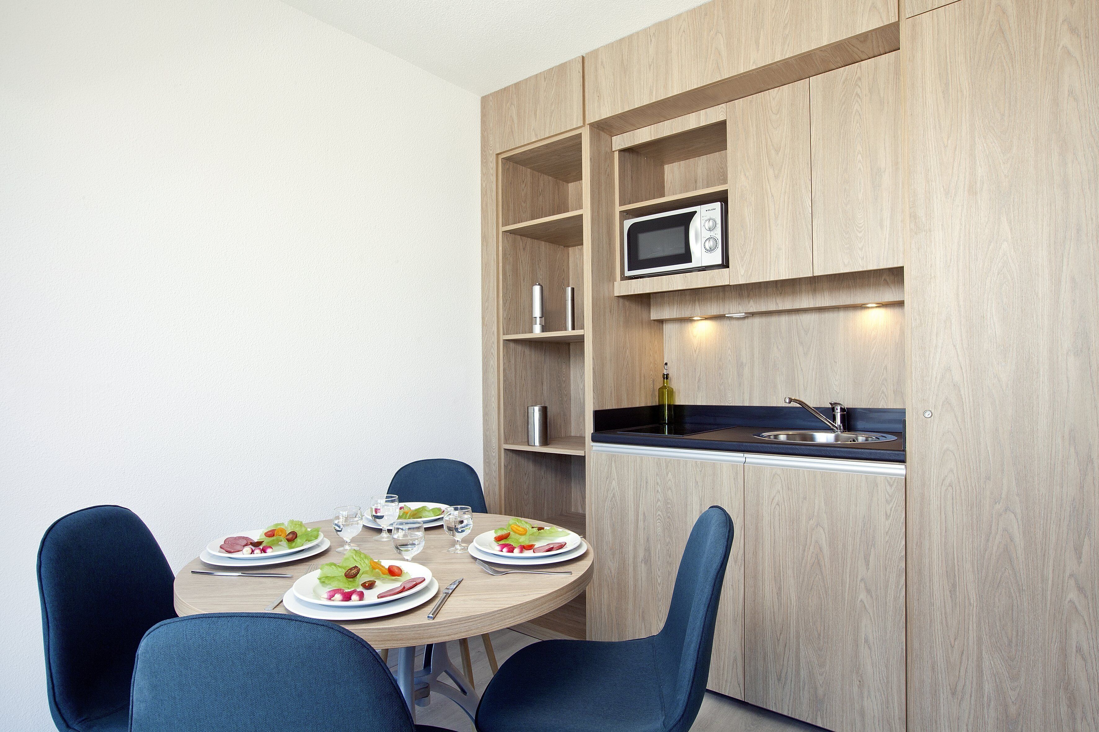 A compact kitchen with a microwave, sink, and wooden cabinets. There is a dining table with chairs and plates of food.