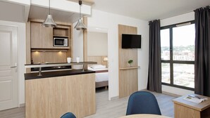 Una habitación de hotel moderna con kitchenette, cama, televisión y un ventanal grande con cortinas.