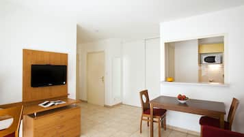 A compact room with a wooden TV stand, a dining table with chairs, a microwave, and a small window.