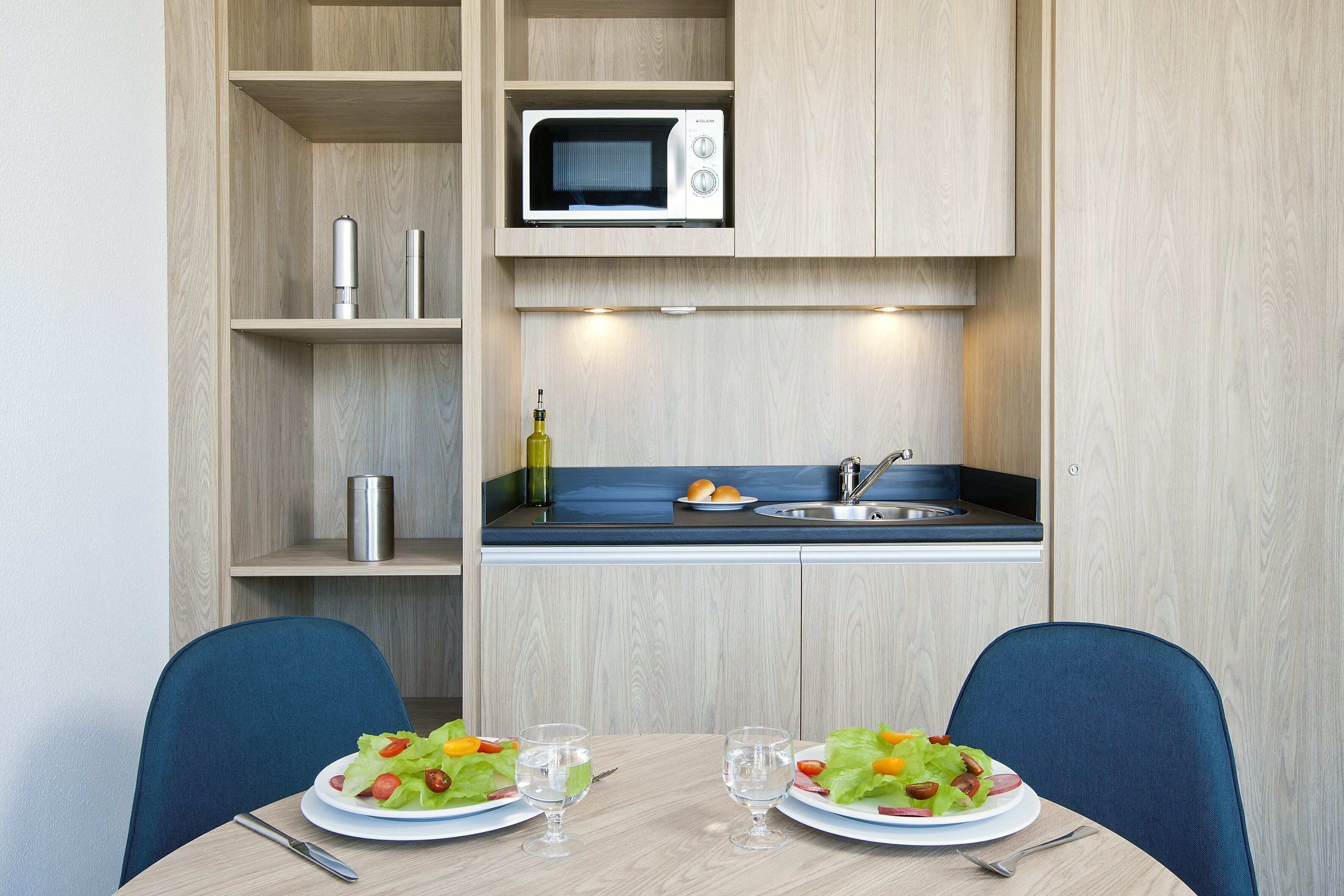 Une petite cuisine équipée d’un four à micro-ondes, d’un évier et d’une petite étagère sur laquelle se trouvent une bouteille et un récipient. On y trouve également une table à manger avec deux assiettes de salade et deux verres.
