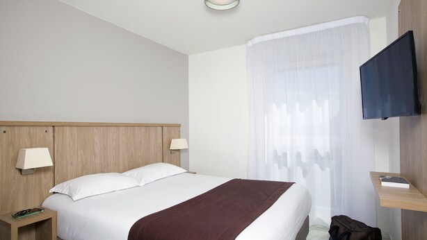 Habitación de hotel con una cama grande, una mesita de noche con lámpara, un televisor montado en la pared y una ventana con cortinas traslúcidas.