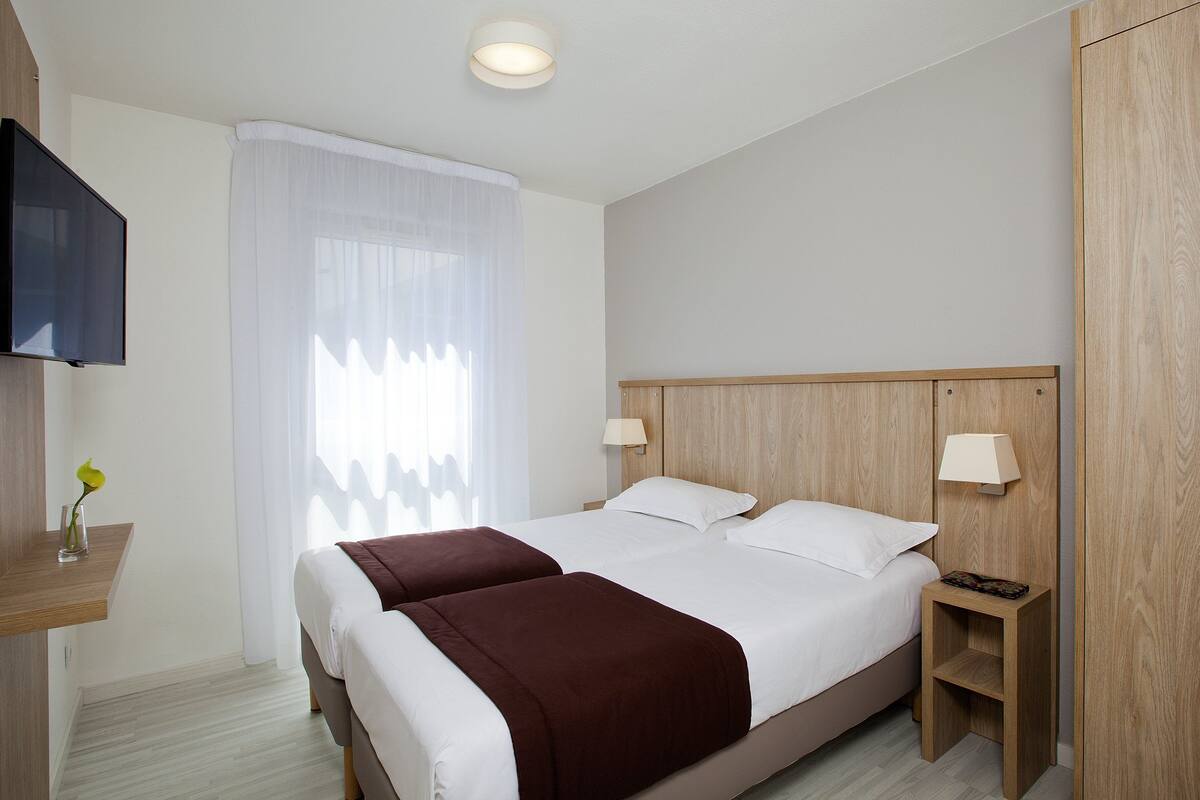 A hotel room with a large bed, two bedside tables, a TV, and a window with sheer curtains.