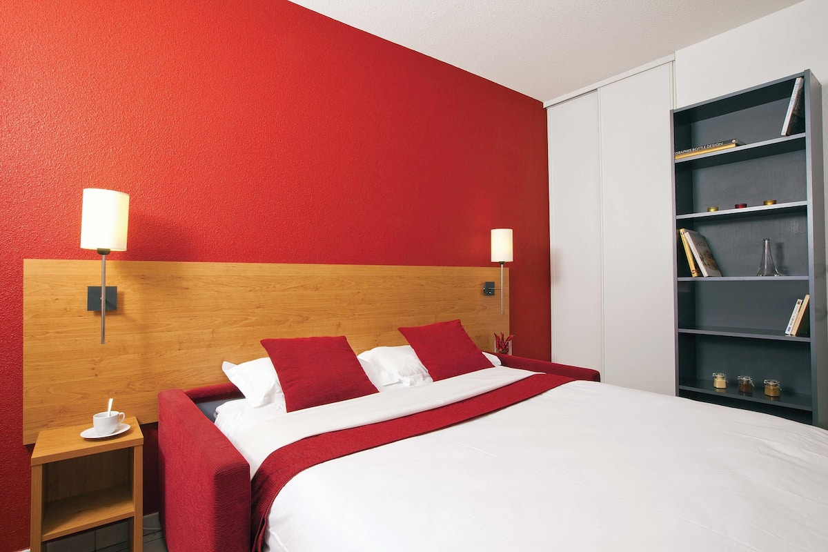 A hotel room with a wooden headboard, a large bed with red and white bedding, two bedside lamps, a small wooden nightstand with a cup, and a bookshelf with books and decorative items.