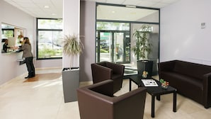 Zona de recepción con recepcionista, mostrador, sillones de cuero marrón, una mesita de centro y plantas en macetas.