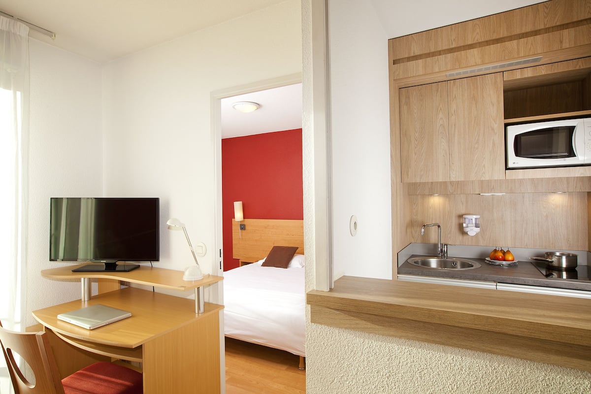 A compact hotel room with a bed, a desk with a laptop, a television, and a kitchenette with a microwave and sink.