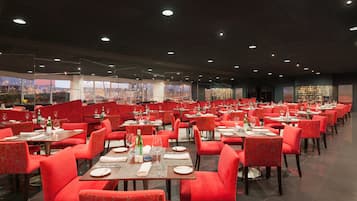 Un restaurante espacioso con sillas y mesas tapizadas en rojo, amplias ventanas que ofrecen vista a la ciudad, y un interior bien iluminado.