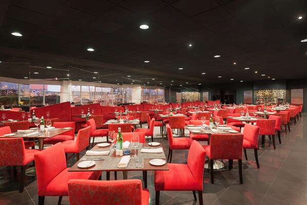 Un restaurante espacioso con sillas y mesas tapizadas en rojo, amplias ventanas que ofrecen vista a la ciudad, y un interior bien iluminado.