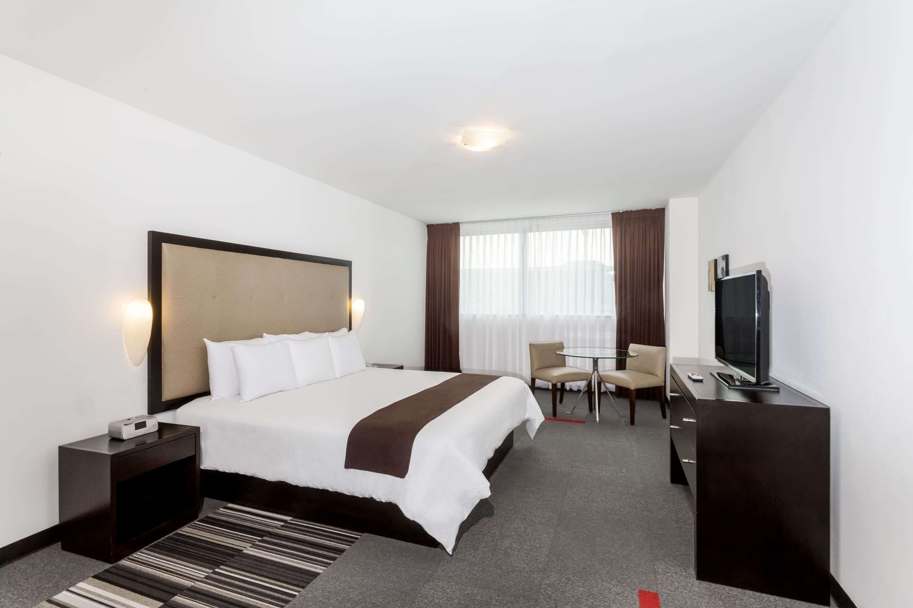Habitación de hotel con cama, mesita de noche, televisión, escritorio, silla y ventana con cortinas.