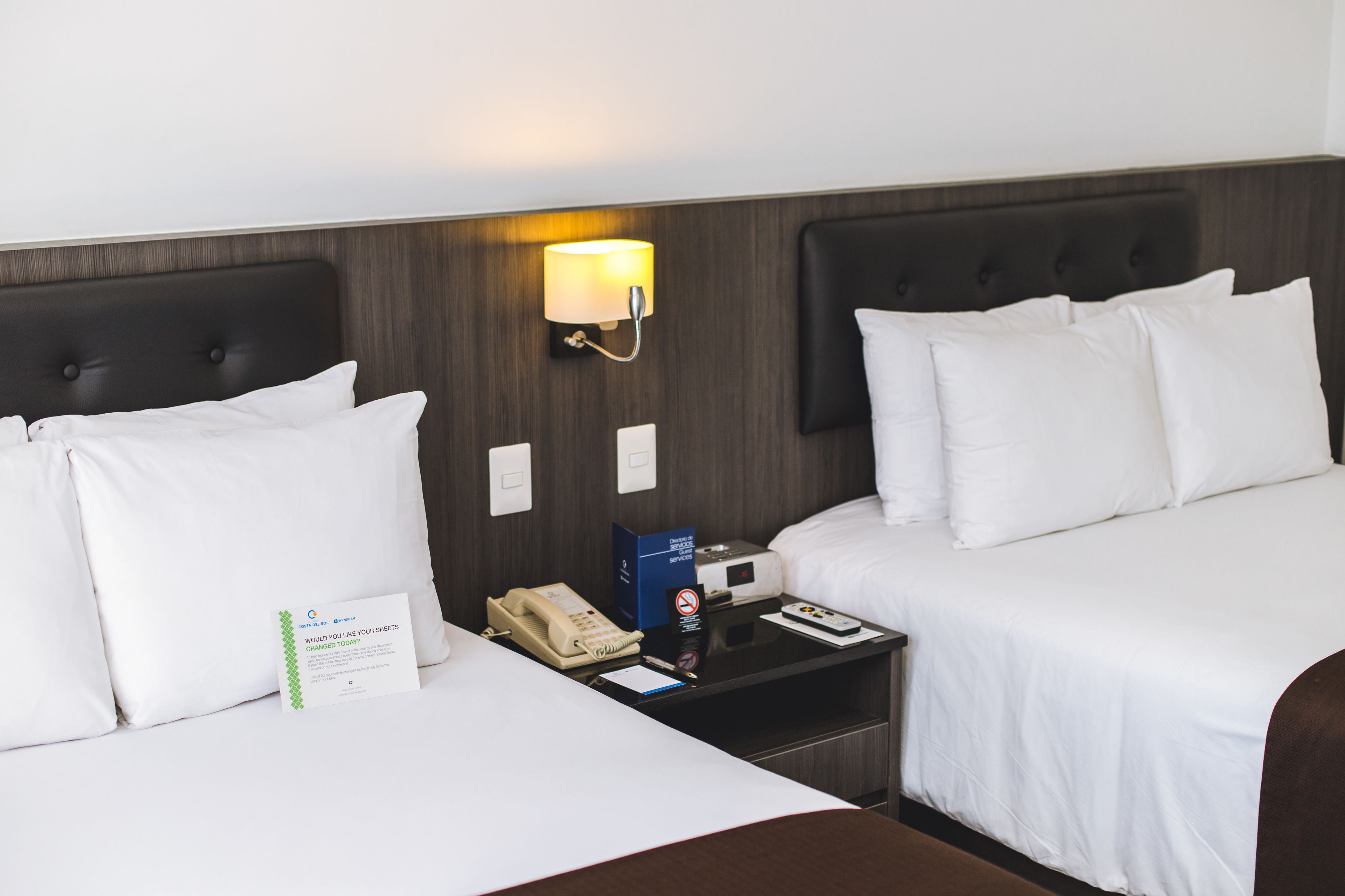 A hotel room with two beds, a nightstand with a lamp, a phone, and a sign.