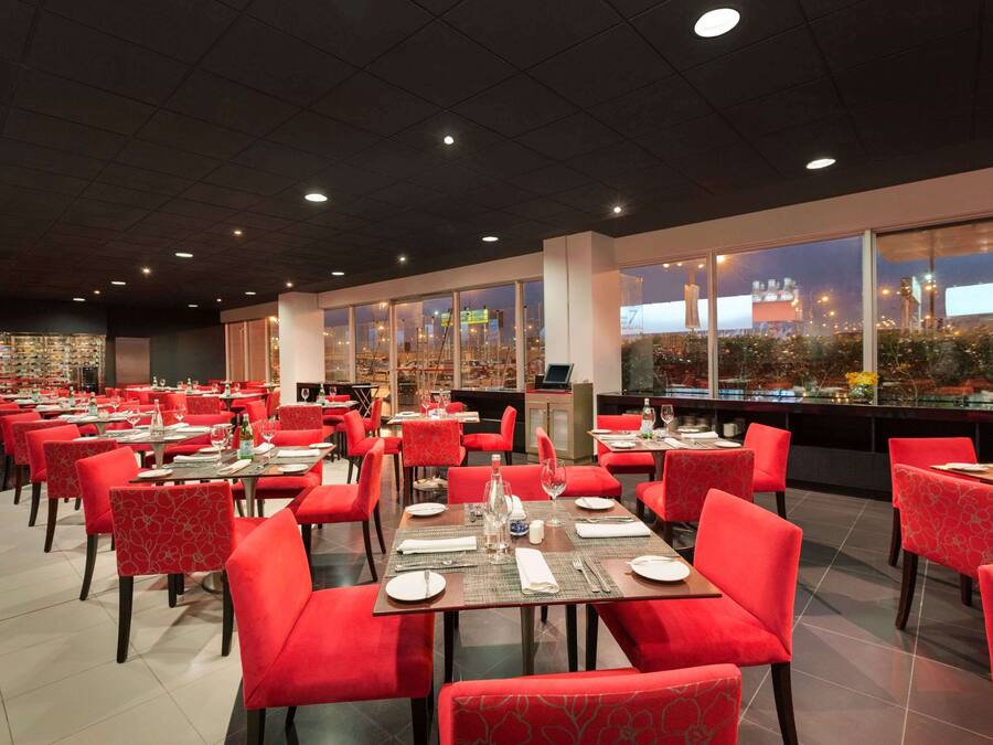 Un restaurante espacioso con sillas y mesas rojas, grandes ventanales que ofrecen vistas del paisaje urbano.