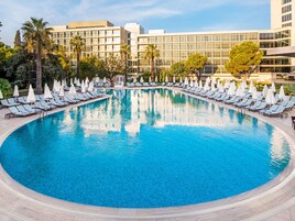 Una piscina amplia y cristalina rodeada de sillones de descanso y sombrillas, con edificios modernos al fondo.
