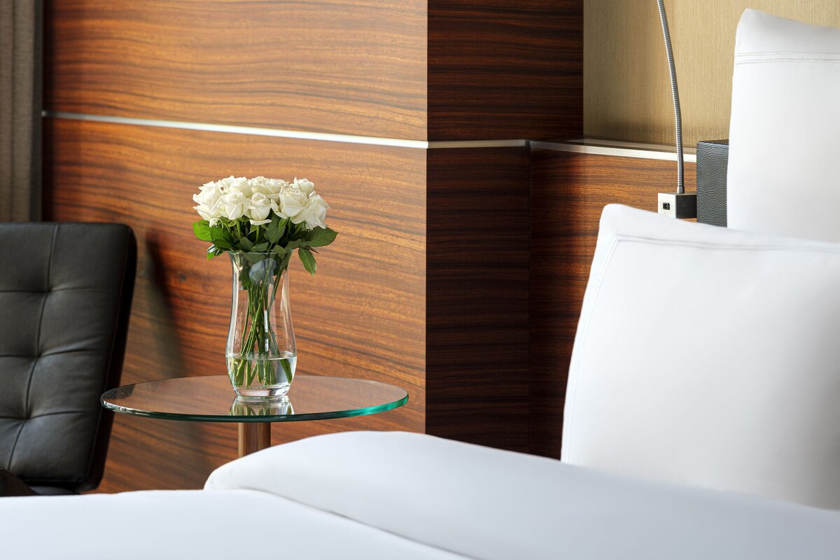 A hotel room with a wooden accent wall, a glass table with a vase of white flowers, a black leather chair, and a white bed.