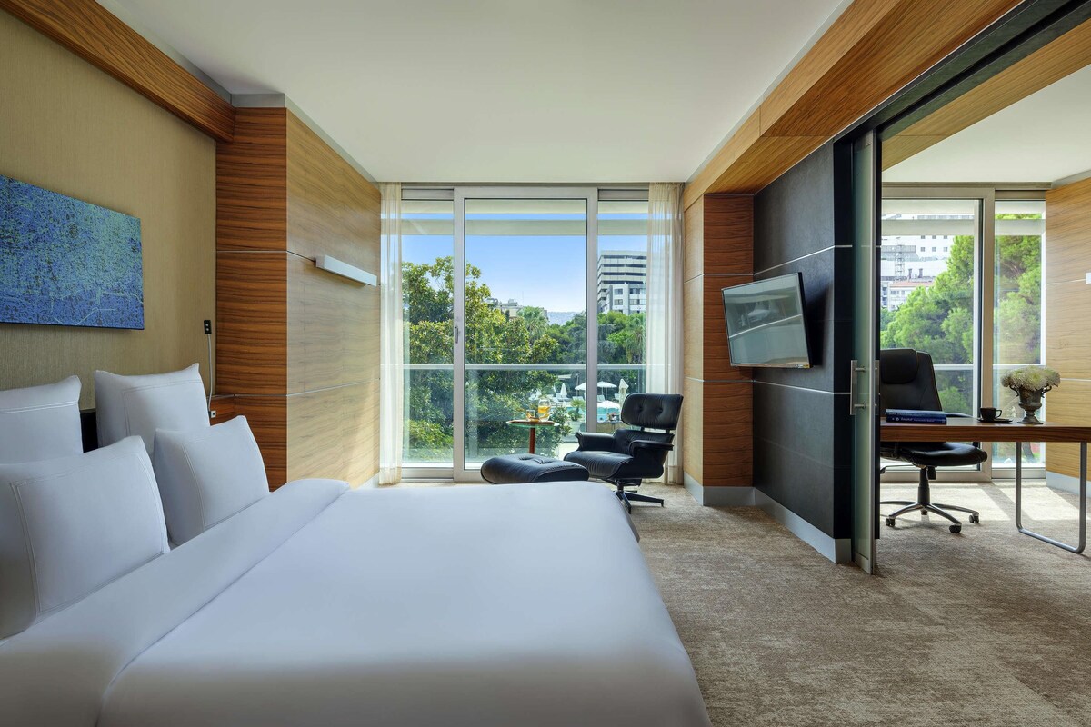 Une chambre d’hôtel avec un grand lit, un bureau avec une chaise, une télévision et une vue sur la verdure et les bâtiments.