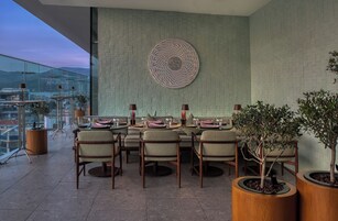 Un comedor con una mesa dispuesta para seis personas, rodeada de sillas, una pieza decorativa en la pared superior y plantas en macetas a cada lado.
